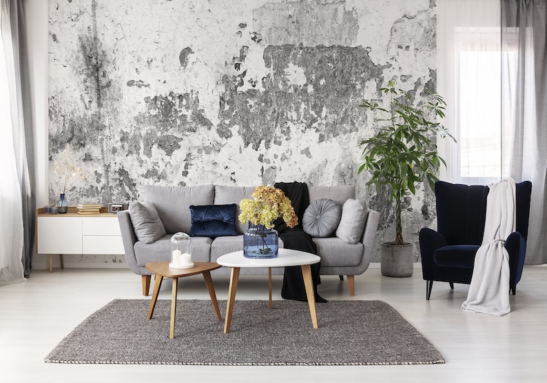 Puede incluir: Un sof&aacute; gris con almohadas azules y grises se encuentra frente a una pared blanca desgastada. Una planta en maceta y un jarr&oacute;n azul con flores amarillas est&aacute;n sobre una mesa de caf&eacute; blanca. Una alfombra gris est&aacute; en el suelo. Un sill&oacute;n azul con una manta blanca est&aacute; en la esquina.