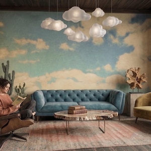 May include: A living room featuring a blue tufted sofa, a mustard yellow chaise lounge, a brown leather armchair, and a live-edge wood coffee table. Above, several white cloud-shaped pendant lights hang against a wall mural of a blue sky.