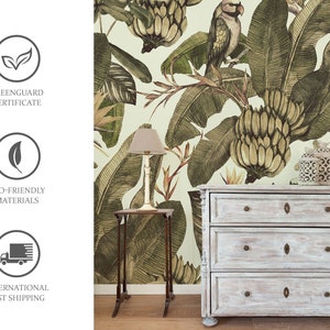 Puede incluir: Una c&oacute;moda de estilo vintage con tres cajones, pintada en un acabado blanqueado. La c&oacute;moda est&aacute; delante de un papel tapiz con estampado tropical que presenta grandes hojas verdes, racimos de pl&aacute;tanos y un loro.