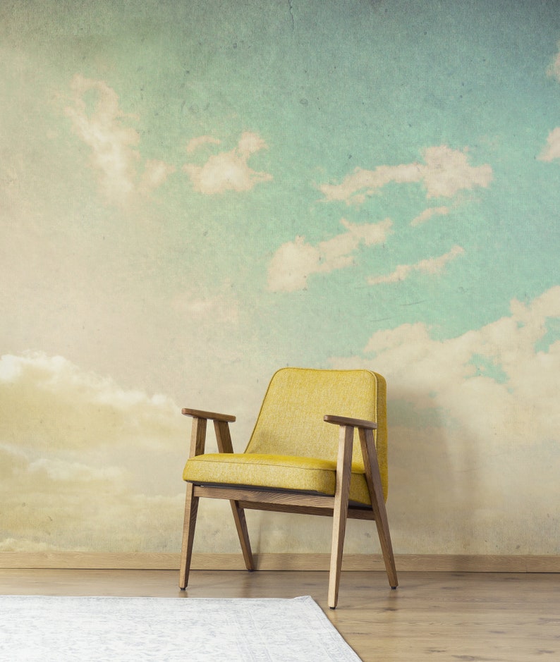 May include: A yellow armchair with wooden arms and legs sits in front of a wall mural depicting a sky with clouds. A white rug is on the wooden floor in front of the chair. The chair has a mid-century modern design.