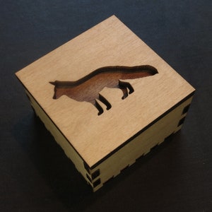 May include: A wooden box with a fox silhouette cutout on the top. The box is made of light brown wood and has a dark brown stain around the edges.
