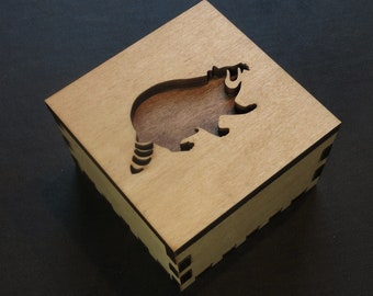 May include: A wooden box with a cutout of a raccoon on the top. The box is made of light brown wood and has a dark brown stain around the edges.