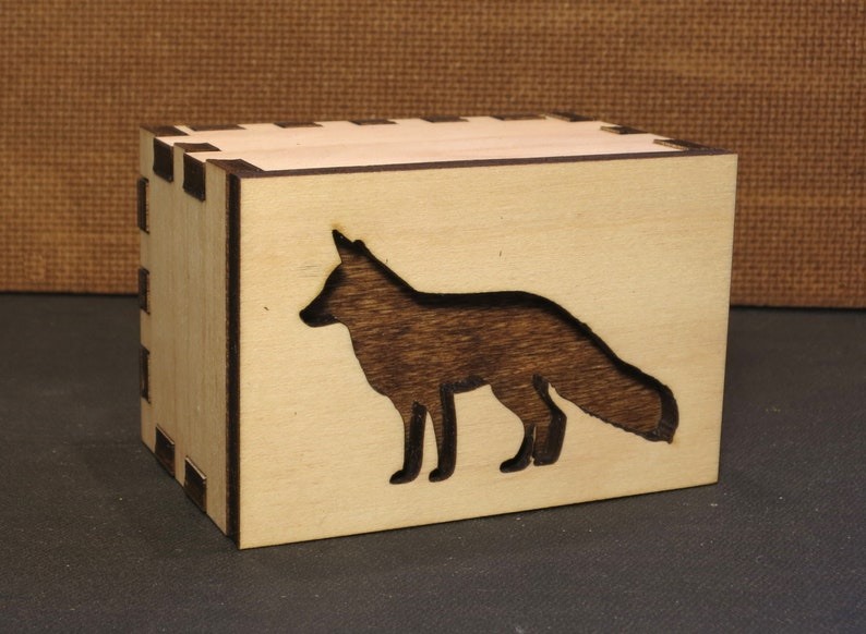 May include: A wooden box with a fox silhouette cutout. The box is made of light wood with a natural finish. The fox is dark brown and is facing to the right.