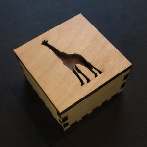 May include: A wooden box with a cutout silhouette of a giraffe. The box is made of light brown wood and has a dark brown stain around the edges.