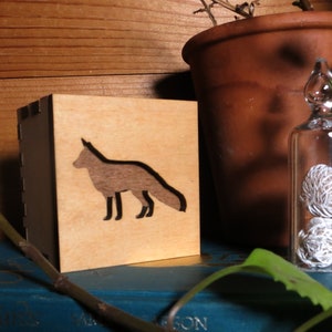 May include: A wooden box with a fox silhouette cutout. The box is sitting on a blue book with a plant in the background.
