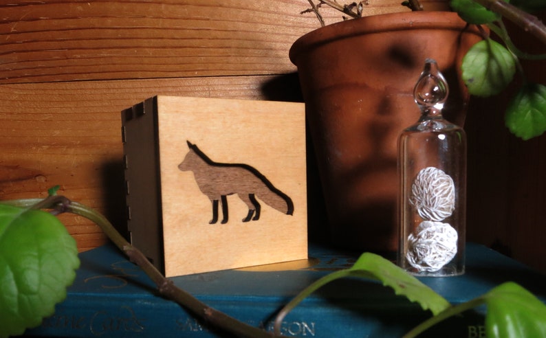 May include: A wooden box with a fox silhouette cutout. The box is sitting on a blue book with a plant in the background.