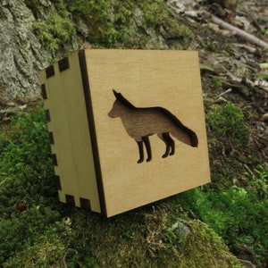 May include: A wooden box with a cutout of a fox silhouette. The box is light brown with a dark brown border. The fox is facing right and has a long tail.