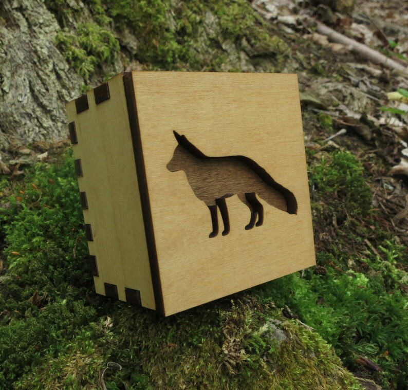 May include: A wooden box with a cutout of a fox silhouette. The box is light brown with a dark brown border. The fox is facing right and has a long tail.