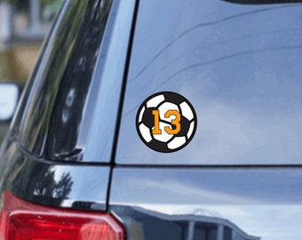 May include: A black car with a soccer ball decal on the back window. The soccer ball is white and black with an orange number 13 in the center.