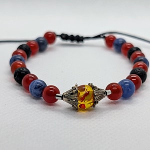 May include: A bracelet with red, blue, and black beads, and a central decorative element. The bracelet has an adjustable black cord. The central bead is yellow and has a silver-coloured metal setting. The bracelet is set against a white background.
