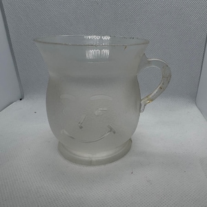 May include: A frosted glass mug with a handle and a smiling face design. The mug has a rounded body and a flared top. The handle is curved and attached to the side. The mug is made of clear glass.