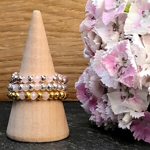 May include: Three stacked rings made of gold, silver, and rose gold beads with pink quartz stones.