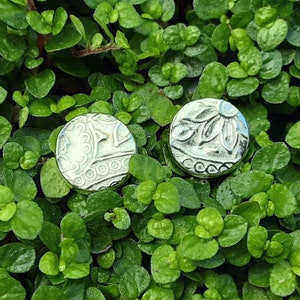 May include: Two silver metal buttons with intricate designs. One button features a floral pattern, while the other has a more geometric design.