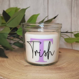 Può includere: Una candela bianca in un barattolo di vetro trasparente con il nome "Trish" in corsivo nero e una "T" viola sul davanti. La candela è su una fetta di legno rotonda con bordo di corteccia naturale. Sullo sfondo ci sono foglie verdi.