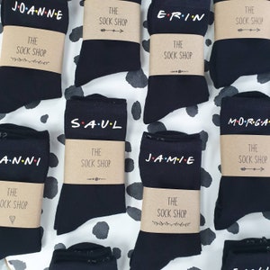 May include: Black socks with names printed in a colourful font, styled after a well-known TV show. Each pair is wrapped with a brown paper band that reads "The Sock Shop". The socks are displayed on a black and white polka dot surface.