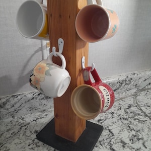 Op de afbeelding: Een houten bekerrek met een zwarte basis houdt vijf bekers vast. De bekers zijn in verschillende kleuren en ontwerpen, waaronder een met het woord "Nana" erop.