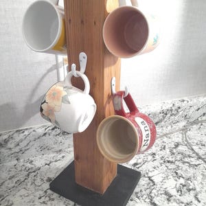 Op de afbeelding: Een houten bekerrek met een zwarte basis. Het rek heeft vijf haken en houdt vier bekers vast. Een beker is rood met het woord "Nana" erop.