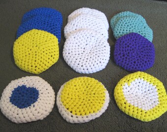 May include: A collection of handmade, crocheted coasters in various colors. The coasters are round and feature a variety of colors, including blue, white, yellow, teal, and purple. Some coasters have contrasting color details.