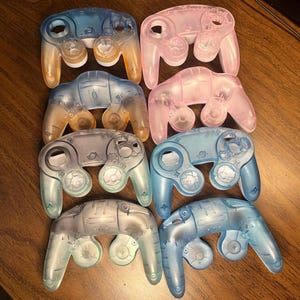 May include: A collection of translucent game controller shells in various colors, including blue, pink, and gray. The controllers have a classic design with buttons and joystick openings. The shells are arranged on a wooden surface.