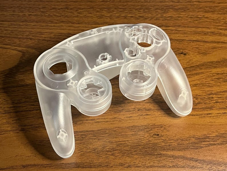 Op de afbeelding: Een doorzichtige plastic behuizing voor een videogamecontroller. De controller heeft een afgeronde vorm met twee handvatten, een D-pad, knoppen en een joystickgebied. De behuizing is ongeverfd en lijkt een vervangend onderdeel te zijn.
