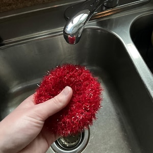 May include: A red knitted dish scrubber in a stainless steel kitchen sink.