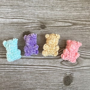 May include: Four small crocheted teddy bears in different colors: light blue, purple, beige, and pink. The bears are arranged in a row on a wooden surface.