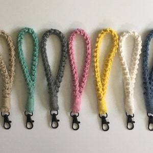 May include: A collection of seven braided wrist lanyards in various colors, including beige, teal, gray, pink, yellow, off-white, and blue. Each lanyard features a black metal clasp for attaching keys or other small items. The lanyards are arranged in a row against a white background.