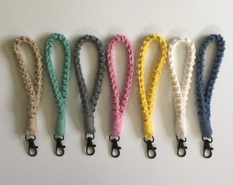 May include: A collection of seven braided wrist lanyards in various colors, including beige, teal, gray, pink, yellow, off-white, and blue. Each lanyard features a black metal clasp for attaching keys or other small items. The lanyards are arranged in a row against a white background.