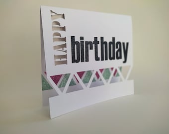 May include: A white card with the words "Happy birthday" cut out. The card has a decorative border of pink and green glitter triangles.