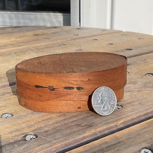 May include: A small, oval-shaped wooden box with a reddish-brown finish. The box is made of thin strips of wood and has a lid. A quarter coin is next to the box for size comparison. The box is sitting on a wooden surface.