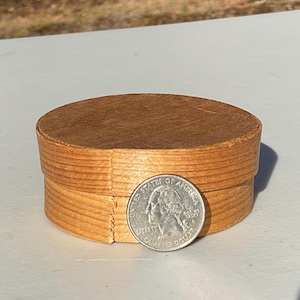 May include: A small, round, wooden box with a lid. The box is made of thin, light brown wood strips. A U.S. quarter coin is next to the box for size comparison. The box is sitting on a white surface.