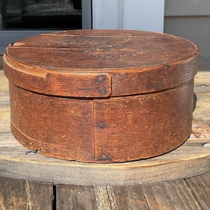 May include: A circular, antique wooden box with a lid. The box is a warm brown colour and appears to be made of wood. The box is sitting on a wooden spool. The box is likely used for storage.