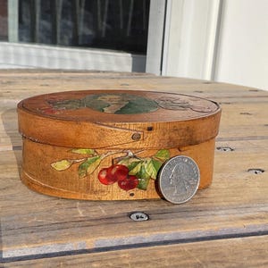 May include: Oval wooden box with a lid featuring a painted design. The box is decorated with a cherry branch and red cherries. A silver coin is placed next to the box for size comparison. The box is sitting on a wooden surface.