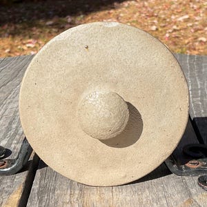 Peut inclure: Couvercle en céramique beige avec un bouton arrondi, posé sur une surface en bois. Le couvercle est circulaire et a une surface texturée. Le couvercle est soutenu par deux supports décoratifs en fer forgé.