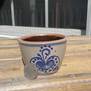 以下が含まれることがあります： 内側が濃い茶色で、青いハートのデザインが施された、小さなベージュ色の陶器のボウル。 ボウルの直径は約7.6cm、高さは5cmです。 サイズ比較のためにボウルの横にコインがあります。 ボウルは木製の表面に置かれています。