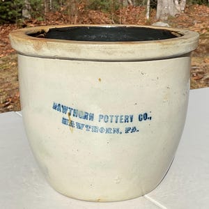 May include: A vintage, off-white ceramic Hawthorn Pottery Co. crock with a dark interior. The crock has a rounded body and a wide, light brown rim. Blue text on the front reads "HAWTHORN POTTERY CO., HAWTHORN, PA."