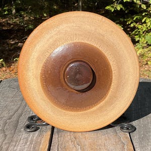 May include: A handmade, round ceramic bowl with a light brown exterior and a dark brown interior. The bowl has a circular design with a central, dark brown, rounded feature. It sits on a decorative, wrought iron stand.
