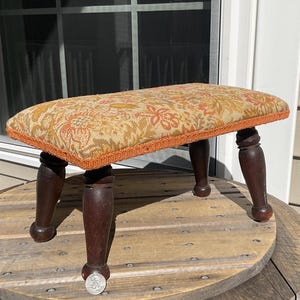 May include: A small, rectangular footstool with a floral patterned cushion in shades of orange, yellow, and green. The cushion is trimmed with orange piping. The dark brown wooden legs are turned and have a rounded foot.
