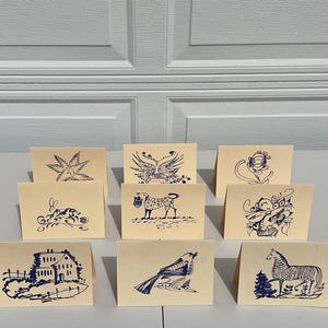 May include: A collection of ten cream-colored cards, each featuring a unique blue ink illustration. The drawings depict various subjects, including a star, bird, and horse. The cards are arranged on a white surface.