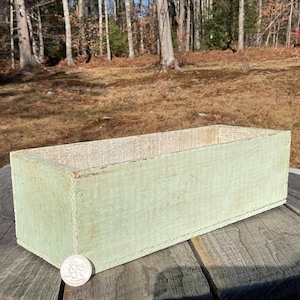 May include: A rectangular wooden planter box with a light green painted finish. The box is empty and sits on a weathered wooden table. A coin is next to the planter. The background shows a natural outdoor setting with trees.