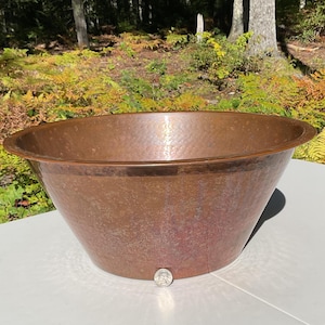 May include: A large, round, hammered copper basin with a wide, flat rim. The exterior surface features a textured, dimpled finish characteristic of hand-hammered metal. A single coin is placed at the base for scale reference.