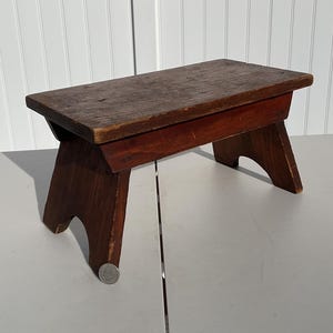 May include: A small, antique wooden step stool with a rectangular top and angled legs. The stool is stained a dark reddish-brown color, showing the wood grain. The stool is approximately 12 inches long, 8 inches wide, and 8 inches tall.