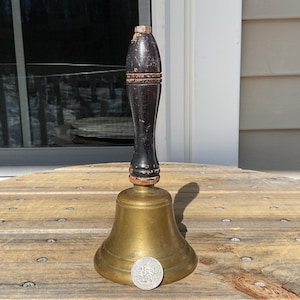 May include: A brass bell with a dark wooden handle. The bell has a flared base and a small coin is placed in front of it. The handle is dark brown and black with a textured appearance. The bell is sitting on a wooden surface.