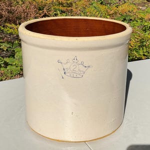 Peut inclure: Un pot en grès cylindrique blanc cassé avec un bord épais et un intérieur brun foncé. L'extérieur présente un motif bleu estampillé représentant une couronne avec le chiffre 2 et les lettres U.S.A. centrés sur la face avant du récipient.