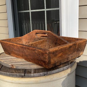 May include: A rustic, rectangular wooden tray with a handle, divided into three sections. The tray is made of weathered, dark brown wood, showing signs of age and use. It sits on a round, wooden spool.