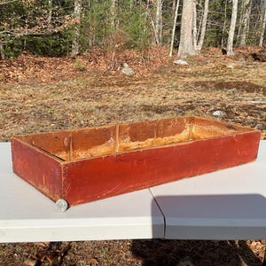 May include: A rectangular, wooden box with a weathered, red exterior. The box is open at the top and appears to be handmade. The interior shows the natural wood grain. A coin is placed on the table next to the box.