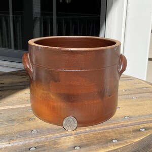 以下が含まれることがあります： 両側に取っ手が付いた茶色の陶器製の壺。壺は円筒形で、縁がわずかにフレアしており、中央にバンドがあります。スケールとして壺の隣にコインがあります。壺は木製のスプールに置かれています。