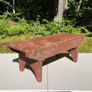 May include: A rectangular wooden bench with a distressed reddish-brown finish. The bench features a flat top surface and four sturdy legs with arched cutouts at the base. The wood shows visible signs of wear and texture across the entire surface.
