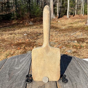 May include: A vintage, light-coloured wooden butter paddle with a long handle and a rectangular paddle. The paddle is resting on a weathered wooden surface with decorative metal accents. A coin rests at the base of the paddle.