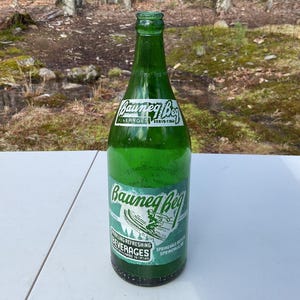 May include: A vintage green glass Bauneg Beg Beverages bottle. The label features a skier graphic and the words "Refreshing Beverages." The bottle is on a white surface with a forest background.